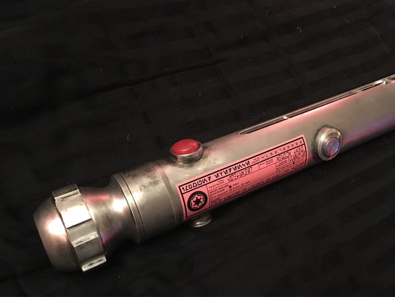May include: A silver lightsaber hilt with a red button and a black and white sticker with the text "120007  KYU7F1M1M  V02-53627".