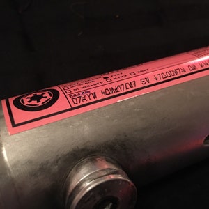 May include: A pink label with a black Imperial symbol and text that reads "D7KYM 4D01K7C/7 4E V7D0007N 0N 1WM", attached to a silver metal tube.