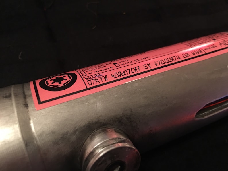 May include: A pink label with a black Imperial symbol and text that reads "D7KYM 4D01K7C/7 4E V7D0007N 0N 1WM", attached to a silver metal tube.