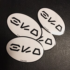 Puede incluir: Cuatro pegatinas ovaladas blancas con la palabra "EVO" impresa en negro en cada pegatina.