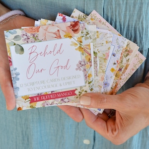 May include: A collection of scripture cards with floral designs, held in a person's hands. The cards feature the text "Behold Our God" and are designed to encourage and uplift. The cards have a variety of floral patterns.