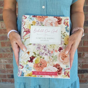 May include: A spiral-bound journal with a floral cover featuring roses and other flowers in shades of pink, peach, and yellow. The journal has the text "Behold Our God" and "Scripture Writing Journal" on the cover. The bottom of the journal has the text "The Ruffled Mango".