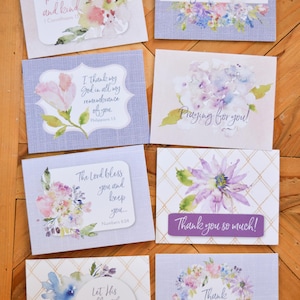 May include: Set of 8 greeting cards with watercolor floral designs and inspirational quotes. Each card features a different floral design and a Bible verse. The cards are printed on a textured paper with a light purple background.