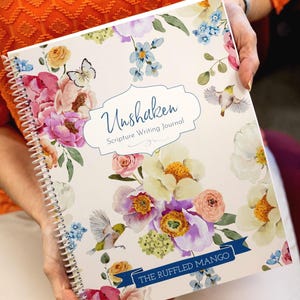 May include: A spiral-bound journal with a floral design featuring pink, purple, and yellow flowers, butterflies, and birds. The journal cover has the text "Unshaken Scripture Writing Journal" and "The Ruffled Mango" on a blue banner.