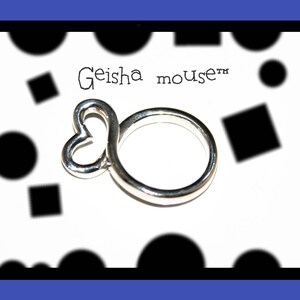May include: A silver ring with a heart-shaped design. The ring is on a white background with black circles and squares. The text "Geisha mouse&trade;" is written on the background.