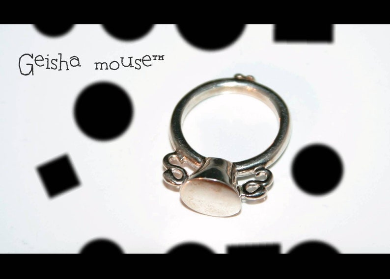 May include: A silver ring with a round, flat top and two small, curved details on either side. The ring is inscribed with the text "Geisha mouse&trade;".