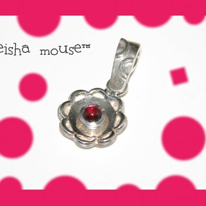 Peut inclure: Un pendentif en argent avec une pierre pr&eacute;cieuse rouge au centre, entour&eacute;e d'un motif floral d&eacute;coratif. Le pendentif est sur un fond blanc avec des pois roses.