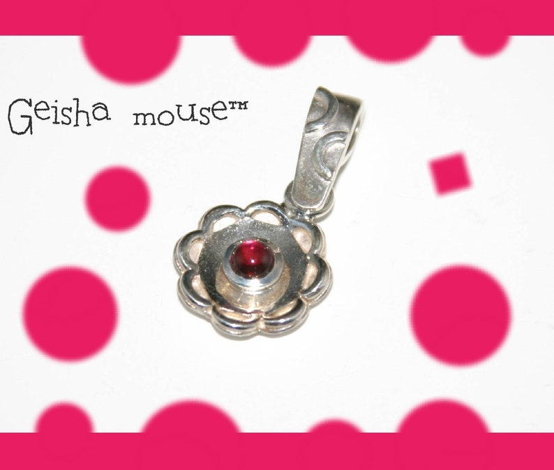 Peut inclure: Un pendentif en argent avec une pierre pr&eacute;cieuse rouge au centre, entour&eacute;e d'un motif floral d&eacute;coratif. Le pendentif est sur un fond blanc avec des pois roses.