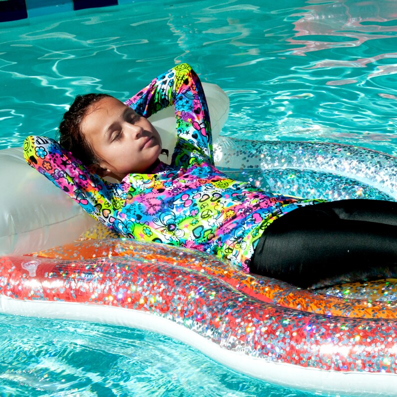May include: A young person wearing a colorful long-sleeved shirt with a black and white pattern floats on a glittery inflatable raft in a swimming pool.