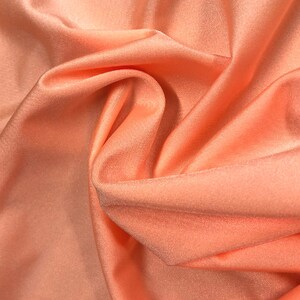 May include: Close-up of a peach-colored fabric with a smooth, silky texture. The fabric is draped and folded, creating soft curves and folds.