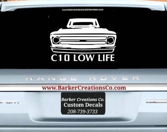 May include: A white silhouette of a C10 pickup truck with the text "C10 LOW LIFE" on a black background. The image is on the back window of a silver Range Rover.