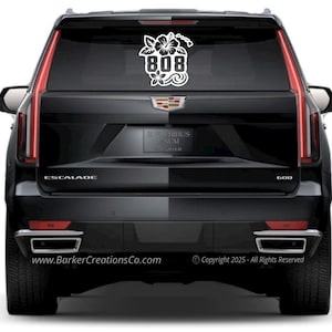 Op de afbeelding: Witte vinyl sticker met het nummer "808" en bloemen- en golfmotieven, aangebracht op de achterruit van een zwarte SUV. De sticker bevat de tekst "BarkerCreationsCo.com" en copyrightgegevens.