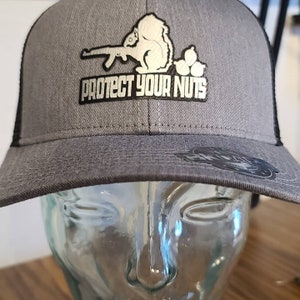 May include: A gray and black trucker hat with a silver embroidered squirrel holding a rifle and the text "Protect Your Nuts".