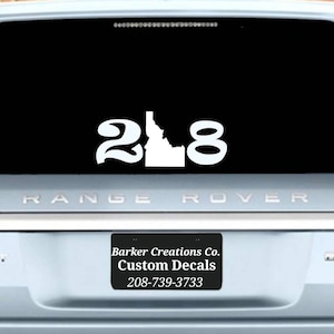 May include: White vinyl decal of the state of Idaho with the numbers 208 on either side. The decal is on the back window of a silver Range Rover.