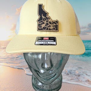 May include: A light yellow trucker hat with a black patch featuring the outline of Idaho and floral details. The hat has a white mesh back and a curved brim. The tag reads "RICHARDSON 115 TRUCKER".