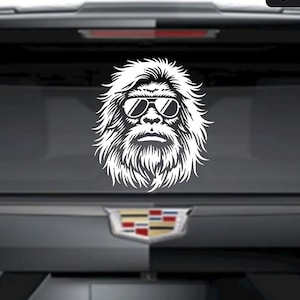 May include: A white vinyl sticker of a cartoon Bigfoot wearing sunglasses. The sticker is on the rear window of a grey car.