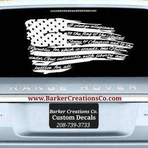 May include: A white vinyl decal of an American flag with the Pledge of Allegiance text. The decal is applied to the back window of a silver Range Rover.