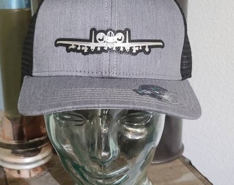 Könnte beinhalten: Eine graue und schwarze Trucker-Kappe mit einem silbernen A-10 Warthog Flugzeug-Motiv. Die Kappe hat ein schwarzes Netzrücken.