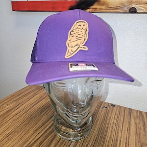 May include: Purple trucker hat with a brown owl patch. The hat has a mesh back and a curved brim.