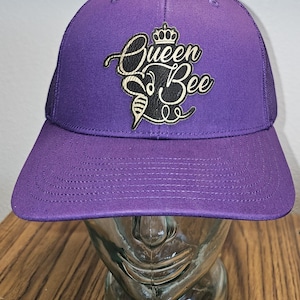 May include: Purple trucker hat with a black and gold embroidered logo that says "Queen Bee" with a crown above the word "Queen".
