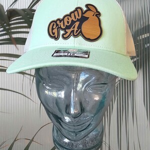 May include: A light green trucker hat with a white mesh back. The hat features the text "Grow A" with a wooden pear graphic. The hat is displayed on a glass head.