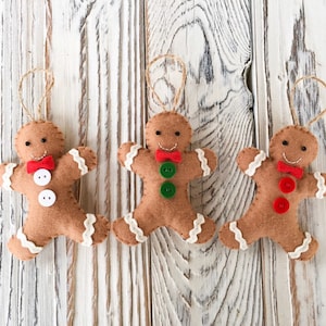 May include: Three felt gingerbread man ornaments with brown bodies, white icing accents, and red or green button details. Each ornament has a red bow tie and a jute string for hanging. The ornaments are on a white wooden background.