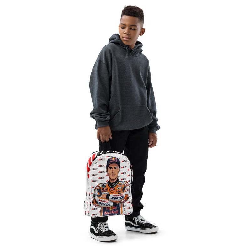 Puede incluir: Una mochila blanca con un estampado gr&aacute;fico de un piloto de motociclismo con un traje de carreras naranja y blanco con el logotipo de Repsol. La mochila tiene un patr&oacute;n repetitivo de logotipos peque&ntilde;os y rayas diagonales rojas y blancas.