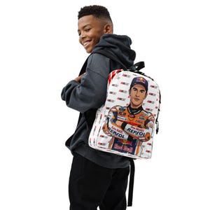 Puede incluir: Una mochila blanca con un patr&oacute;n repetitivo del n&uacute;mero 93 y un gr&aacute;fico central de un piloto de motos con un traje de carreras Repsol naranja, blanco y azul con la marca Red Bull y una gorra a juego, con correas y cremalleras negras.