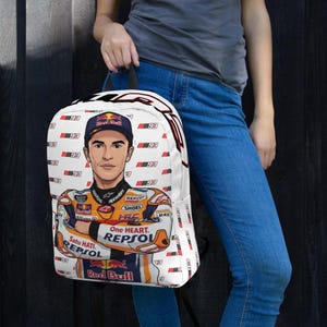 Puede incluir: Una mochila blanca con un patr&oacute;n repetitivo del n&uacute;mero 93 y una ilustraci&oacute;n central de un piloto de motos con un traje de carreras naranja, blanco y azul con logotipos de Red Bull, Repsol y One Heart, adem&aacute;s de una gorra a juego.