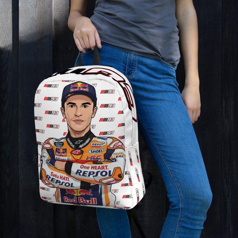 Puede incluir: Una mochila blanca con un patr&oacute;n repetitivo del n&uacute;mero 93 y una ilustraci&oacute;n central de un piloto de motos con un traje de carreras naranja, blanco y azul con logotipos de Red Bull, Repsol y One Heart, adem&aacute;s de una gorra a juego.