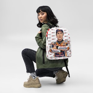 Puede incluir: Una mochila blanca con un patr&oacute;n repetitivo del n&uacute;mero 93 y un gr&aacute;fico grande de un piloto de motos con un traje de carreras de Repsol y Red Bull. El piloto aparece con los brazos cruzados, una gorra y el texto One Heart.