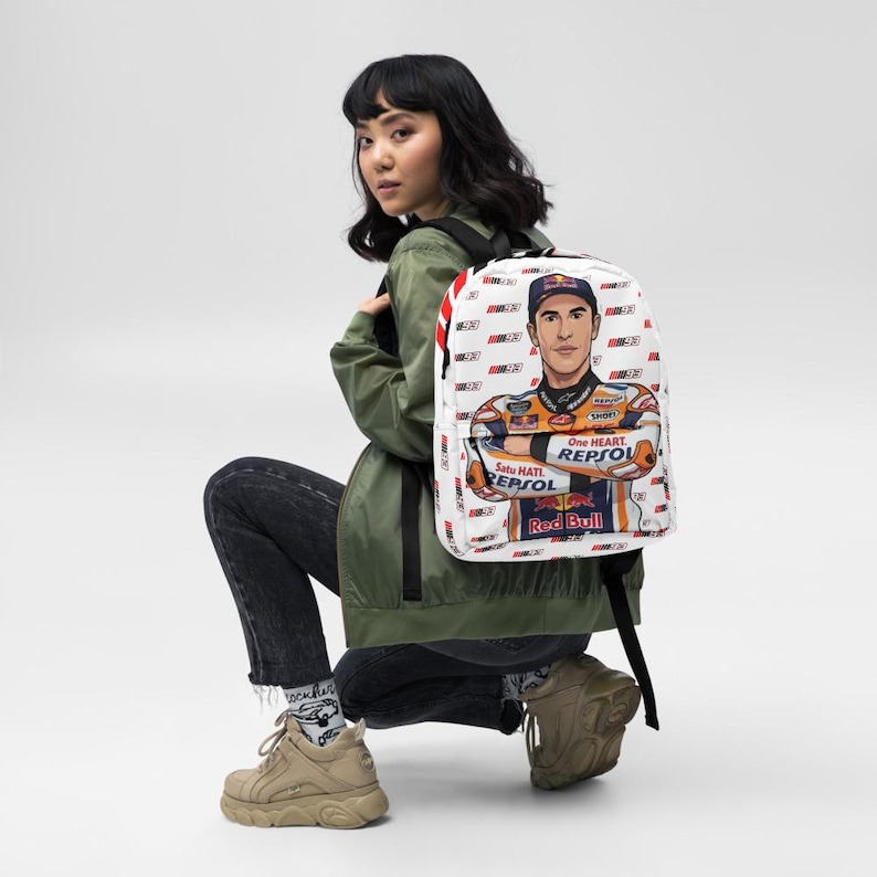 Puede incluir: Una mochila blanca con un patr&oacute;n repetitivo del n&uacute;mero 93 y un gr&aacute;fico grande de un piloto de motos con un traje de carreras de Repsol y Red Bull. El piloto aparece con los brazos cruzados, una gorra y el texto One Heart.