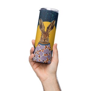 May include: A stainless steel tumbler featuring a whimsical illustration of a hare wearing a floral sweater against a yellow backdrop. The tumbler has a dark blue starry sky background and a silver lid. The design is printed on the tumbler.