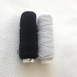May include: Two spools of thread, one black and one white, are shown on a white textured surface.