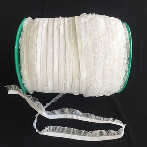 May include: A spool of white elastic trim with a ruffled edge. The trim is wound around a green plastic spool.