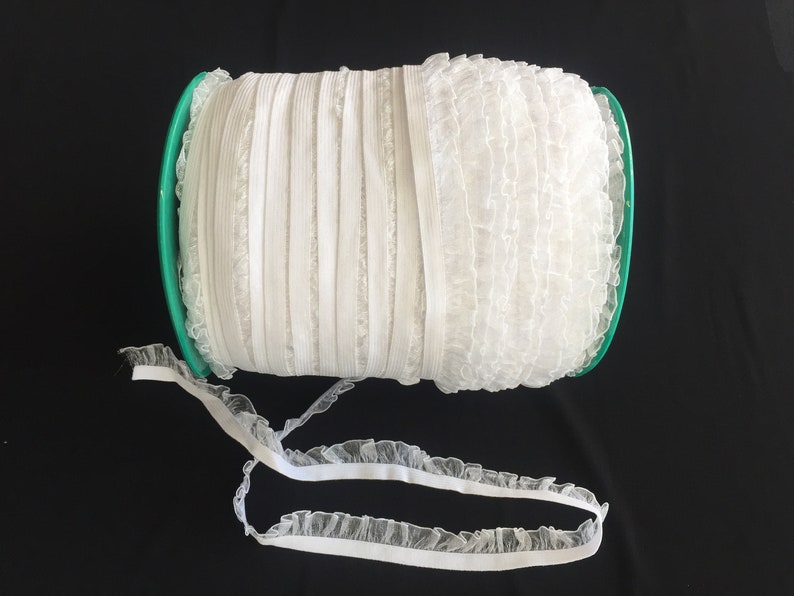 May include: A spool of white elastic trim with a ruffled edge. The trim is wound around a green plastic spool.