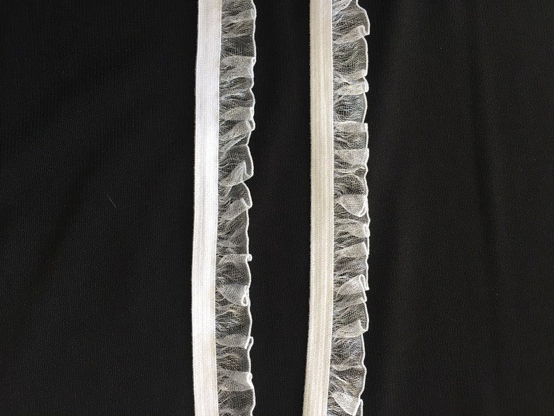 May include: Two white elastic trims with a ruffled tulle edge. The trims are against a black background.