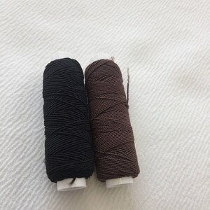 May include: Two spools of thread, one black and one brown, are shown on a white surface.