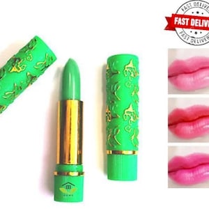 Puede incluir: Barra de labios verde con base dorada y estuche verde decorativo. El estuche está abierto, revelando la barra de labios. La imagen también muestra tres ejemplos del color de la barra de labios en los labios.