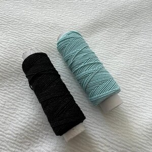 May include: Two spools of thread, one black and one light blue, are on a white textured surface.