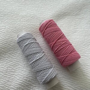 May include: Two spools of thread, one pink and one gray, are on a white textured surface.