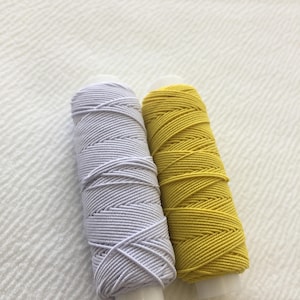 May include: Two spools of thread, one white and one yellow, are wound tightly together. The spools are made of plastic and have a white background.