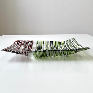 May include: A decorative glass serving dish with a rectangular shape and a wavy design. The dish features vertical stripes in red, white, black, and green. The glass is transparent, allowing light to pass through.