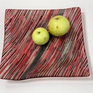 May include: A square glass serving dish with a red, black, and white striped pattern. Two green apples are on the dish.