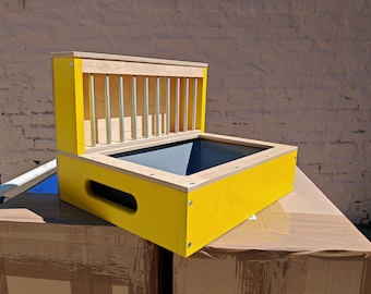 May include: A yellow wooden box with a grey interior and a natural wood top. The box features vertical metal rods on one side. The box is placed on a cardboard box. The box is approximately 40 cm long.