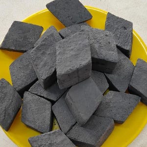 Pode incluir: Um close-up de um monte de briquetes de carvão vegetal preto em uma tigela amarela. Os briquetes têm a forma de diamantes e uma superfície áspera e texturizada.