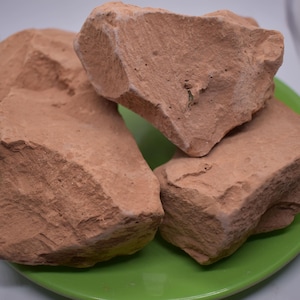 May include: Four large, irregularly shaped pieces of reddish-brown clay on a green plate. The clay is rough and textured.
