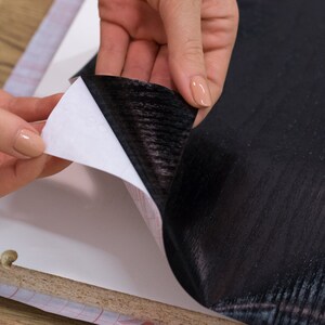 K&ouml;nnte beinhalten: Eine Person klebt eine schwarze Holzmaserung-Kontaktfolie auf eine Oberfl&auml;che. Die Kontaktfolie hat eine wei&szlig;e R&uuml;ckseite, die abgezogen wird, um die Klebeseite freizulegen.