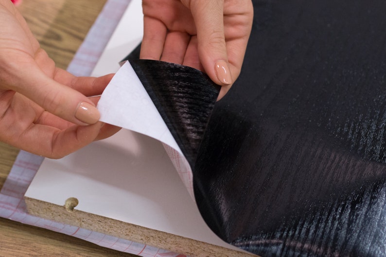 K&ouml;nnte beinhalten: Eine Person klebt eine schwarze Holzmaserung-Kontaktfolie auf eine Oberfl&auml;che. Die Kontaktfolie hat eine wei&szlig;e R&uuml;ckseite, die abgezogen wird, um die Klebeseite freizulegen.