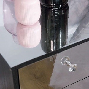 Pu&ograve; includere: Un com&ograve; con specchio con tre cassetti e una manopola di cristallo. Il com&ograve; ha una cornice nera e una superficie riflettente. Due vasi di vetro rosa e un vaso di vetro nero sono sopra il com&ograve;.